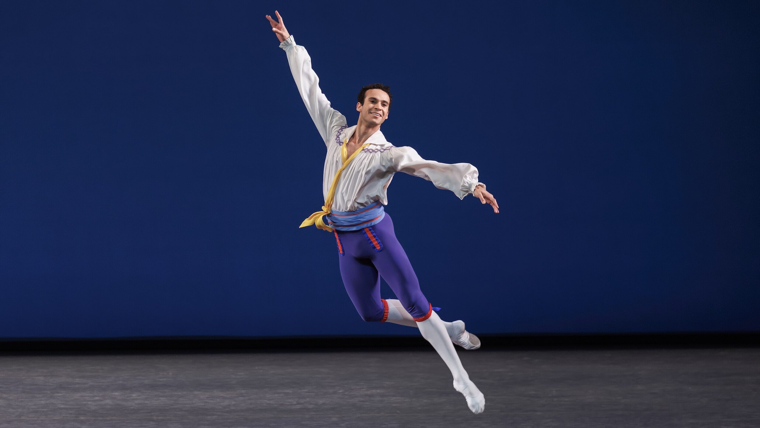 Ryan Tomash does a jeté with his arms extended in a diagonal line. He wears a billowy white blouse, light blue cummerbund, blue knee-length tights over white tights, and white ballet slippers. He performs onstage in front of a blue backdrop.