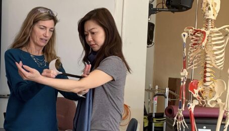 Dr. Linda Bluestein stretches out kinesiology tape to apply to a patient's elbow. The patient, arm lifted with a slight bend, looks down at the tape with concentration.