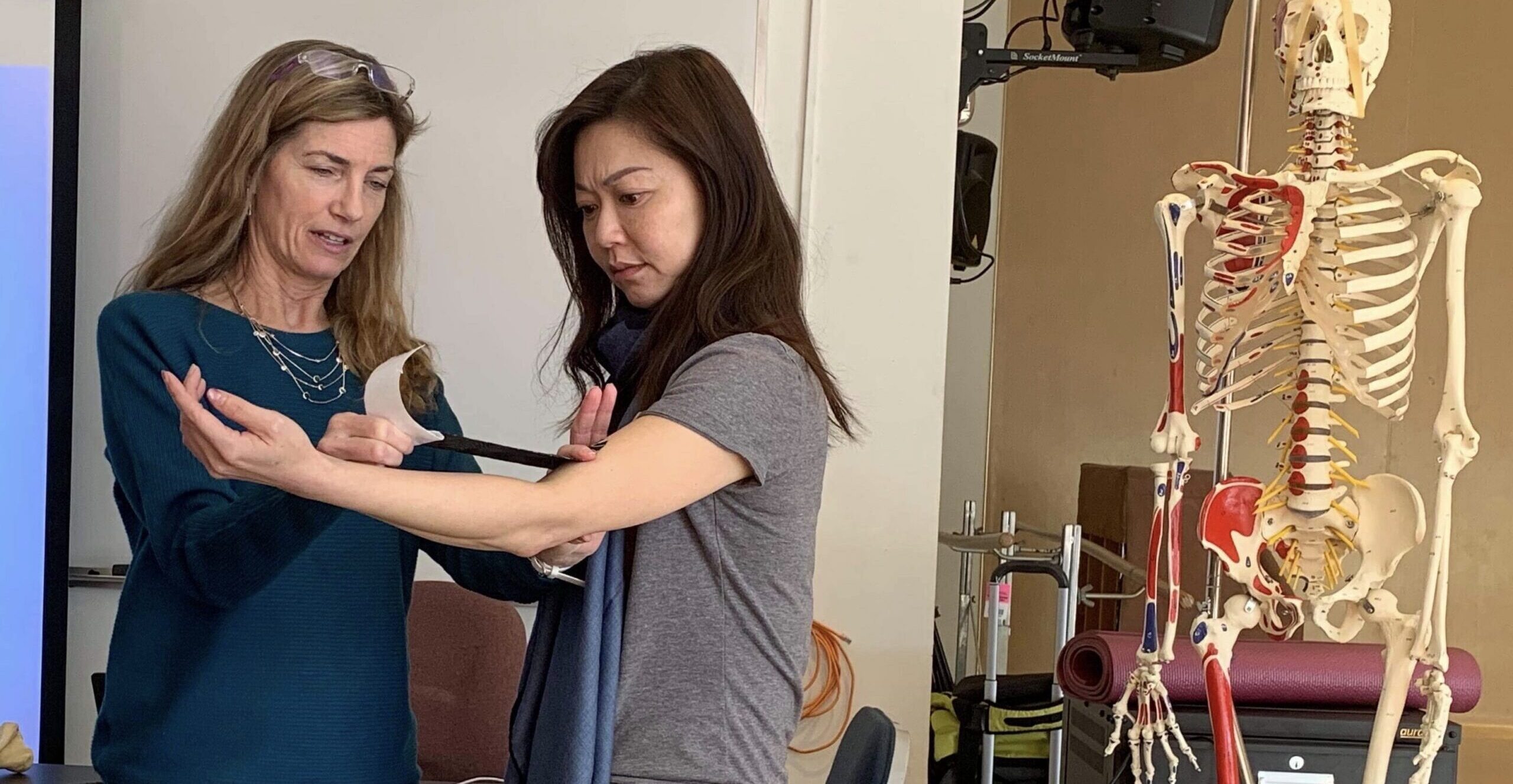 Dr. Linda Bluestein stretches out kinesiology tape to apply to a patient's elbow. The patient, arm lifted with a slight bend, looks down at the tape with concentration.