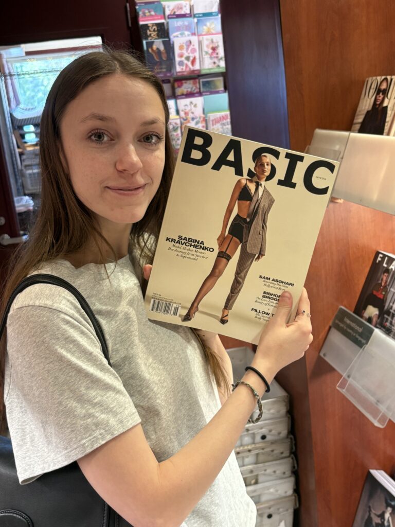 Cassidy Cocke poses in a grey t-shirt and black purse holding a magazine entitled Basic.