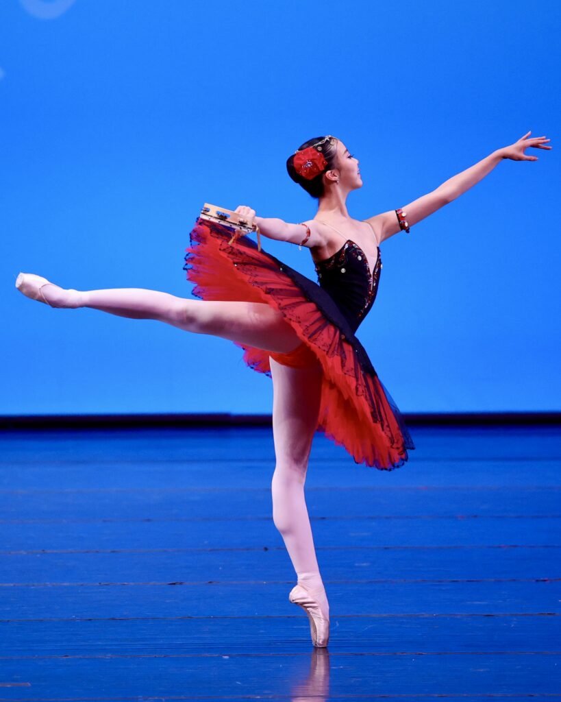 A female dancer stands on pointe in arabesque, holding a tambourine in her back hand. She wears a red and black tutu and a red flower in her dark hair.