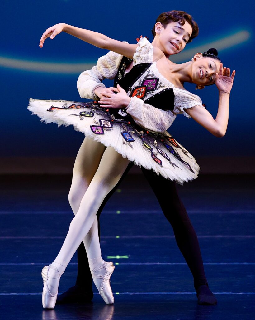 A young boy and girl perform a ballet pas de deux. She stands on pointe and bends backward, her hend to her head, as he stands behind her and holds her by the waist. the girls wears a white tutu with a black bodice with colorful diamond patterns, while her partner wears black tights and a tunic that matches her costume.