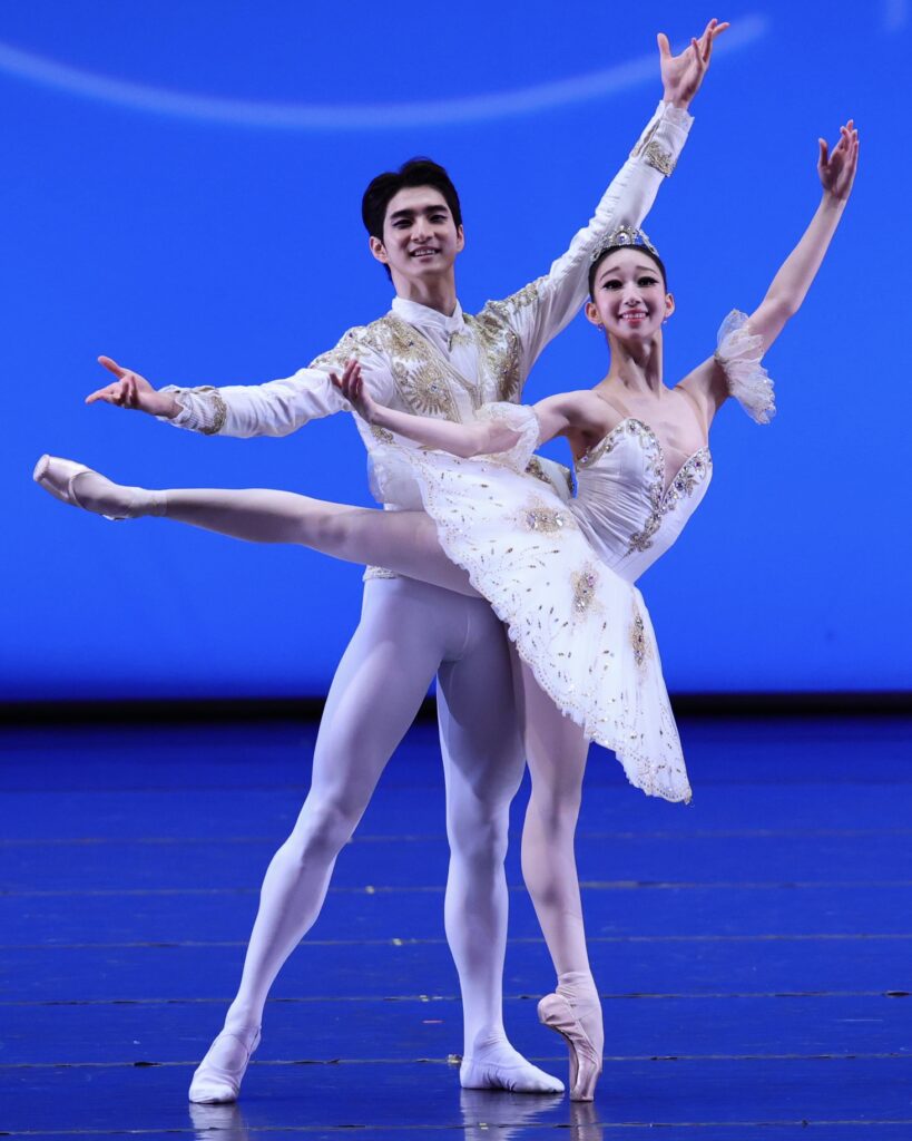 A teenage ballet studen in a white tutu poses in arabesque on pointe, looking out towards the audience. her partner, wearing a white tunic and tights, stand behind her, matching her port de bras.