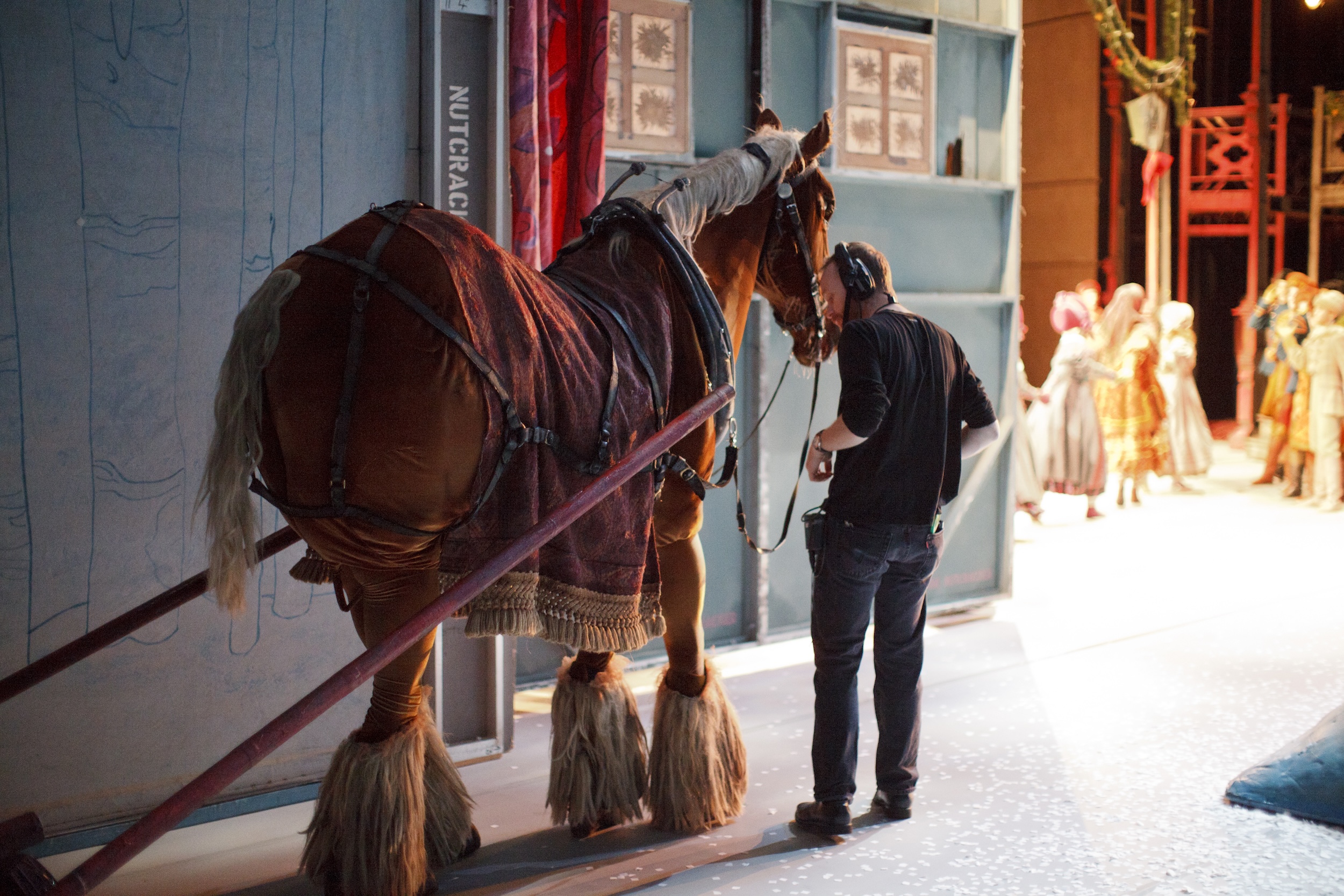 Jeff Morris, wearing a headset, dark sweater, and pants, stands in the wings with two dancers wearing a life-sized horse costume as dancers in costume mill about onstage in the background.