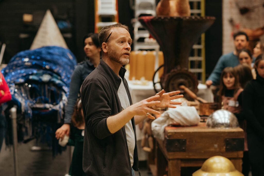 Jeff Morris, wearing a gray t-shirt and a dark gray zip-up sweater, talks to a group of people backstage at a theater during a tour. Props and sets are in the background.