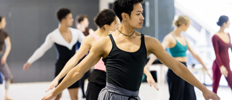 A dancer wearing a black tank top and light grey athletic pants poses in a wide forth position with his arms in a low-V. Behind him are six dancers in the same position.