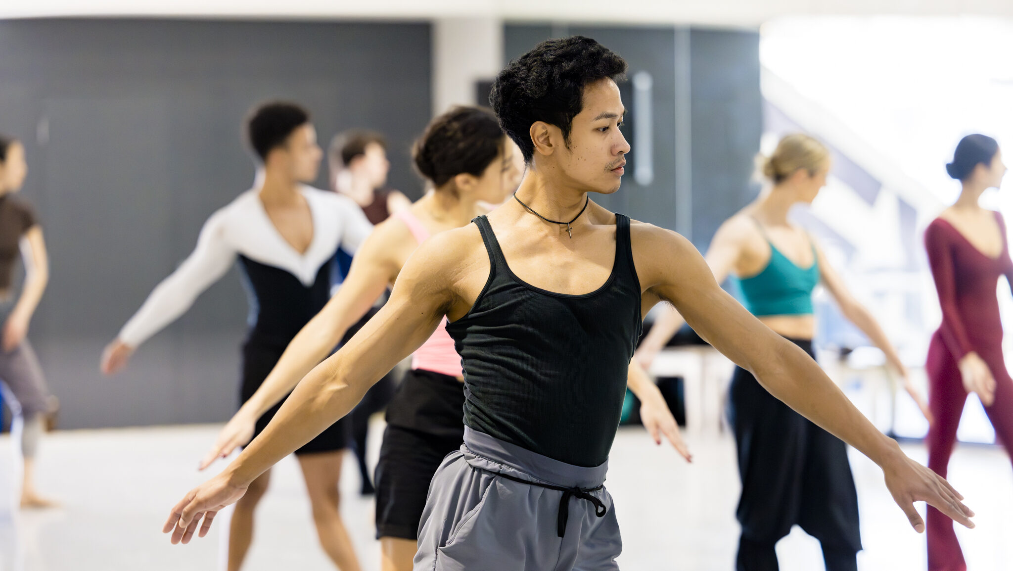 A dancer wearing a black tank top and light grey athletic pants poses in a wide forth position with his arms in a low-V. Behind him are six dancers in the same position.