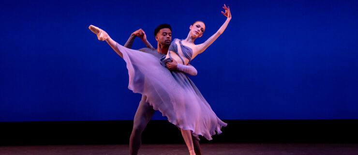 Cassidy Cocke appears on stage in a flowing purple dress and poses on pointe with her right leg extended to the front and her left arm in fifth position. Her partner wears a tight purple unitard and wraps his arm around her to support her position.