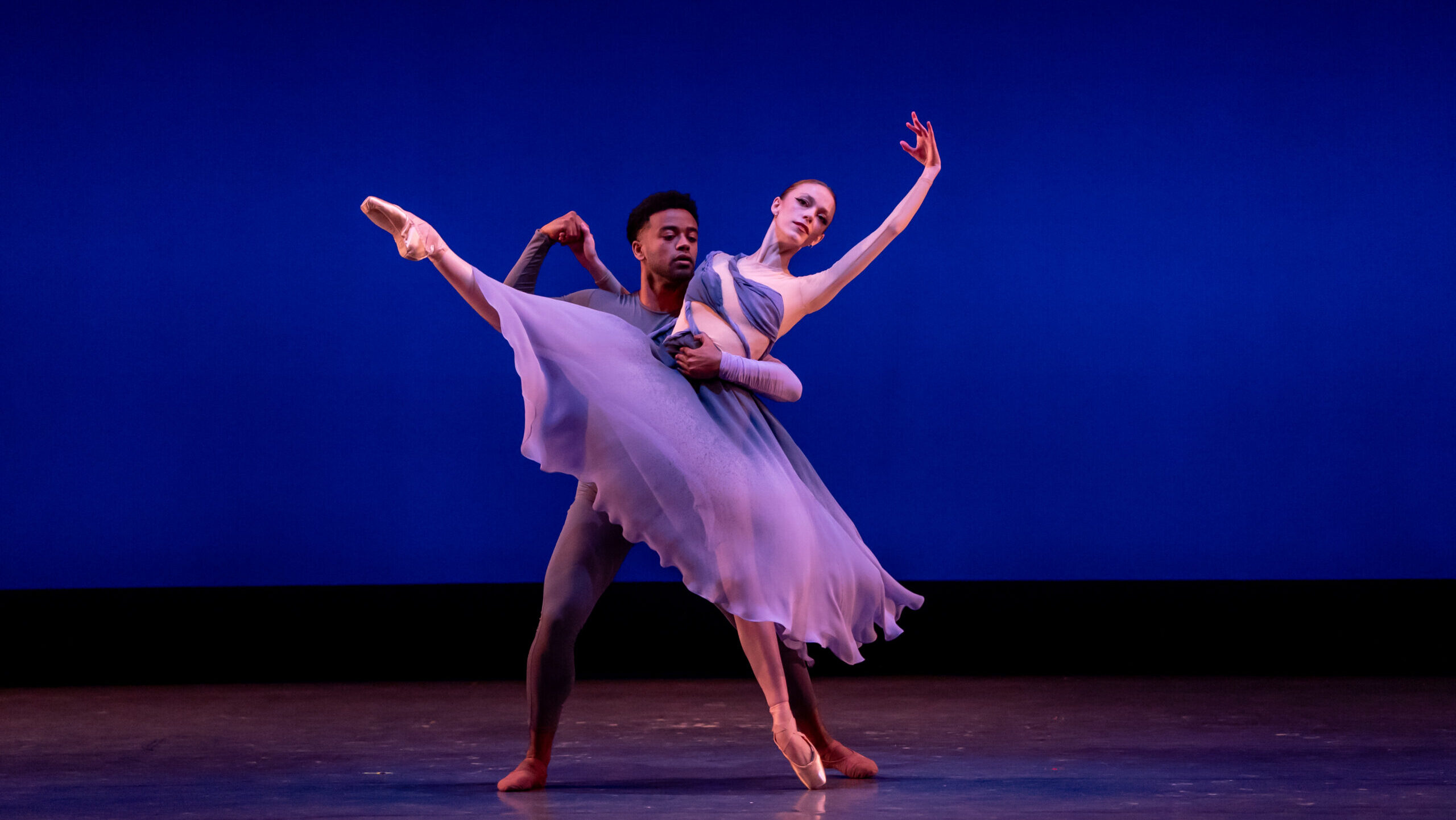 Cassidy Cocke appears on stage in a flowing purple dress and poses on pointe with her right leg extended to the front and her left arm in fifth position. Her partner wears a tight purple unitard and wraps his arm around her to support her position.
