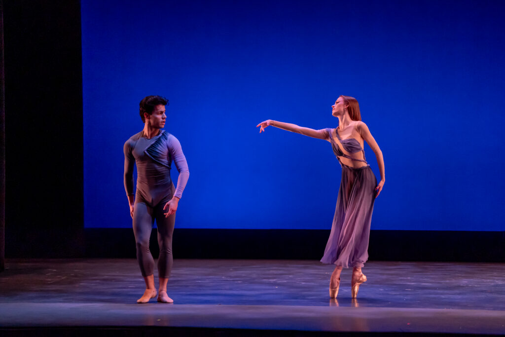 Cassidy Cocke and her partner appear on stage with a solid blue backdrop. She wears a flowing purple dress and stands on pointe in forth position, reaches her right arm toward her partner. He stands looking back toward her.