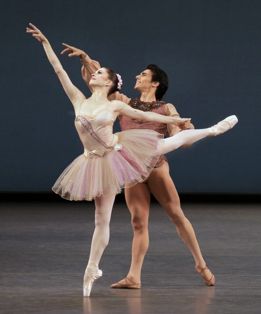 Megan Fairchild, wearing a shirt pink tutu, does a piqué first arabesque on pointe towards stage right. Joaquin de Luz, in a light brown tunic and tights, stands behind her in tendu derriere and holds her left hand.