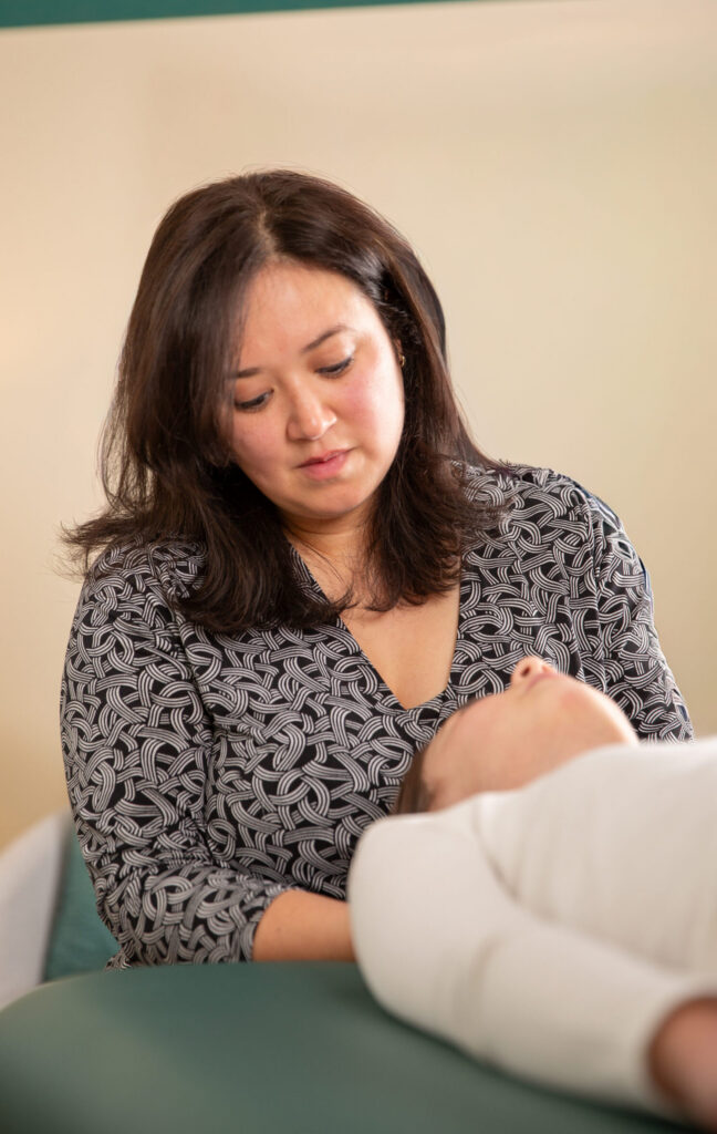 Dr. Aiko Callahan sits at one end of a physical therapy table, her hands resting underneath a patient's head as the patient lies prone on her back.