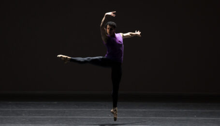 Lasha Khozashvili jumps with one leg extended 90 degrees a la seconde, his arms in high third with a slight twist in his shoulders. He wears black tights and a deep purple tank top.