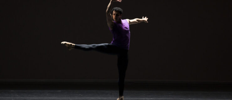 Lasha Khozashvili jumps with one leg extended 90 degrees a la seconde, his arms in high third with a slight twist in his shoulders. He wears black tights and a deep purple tank top.