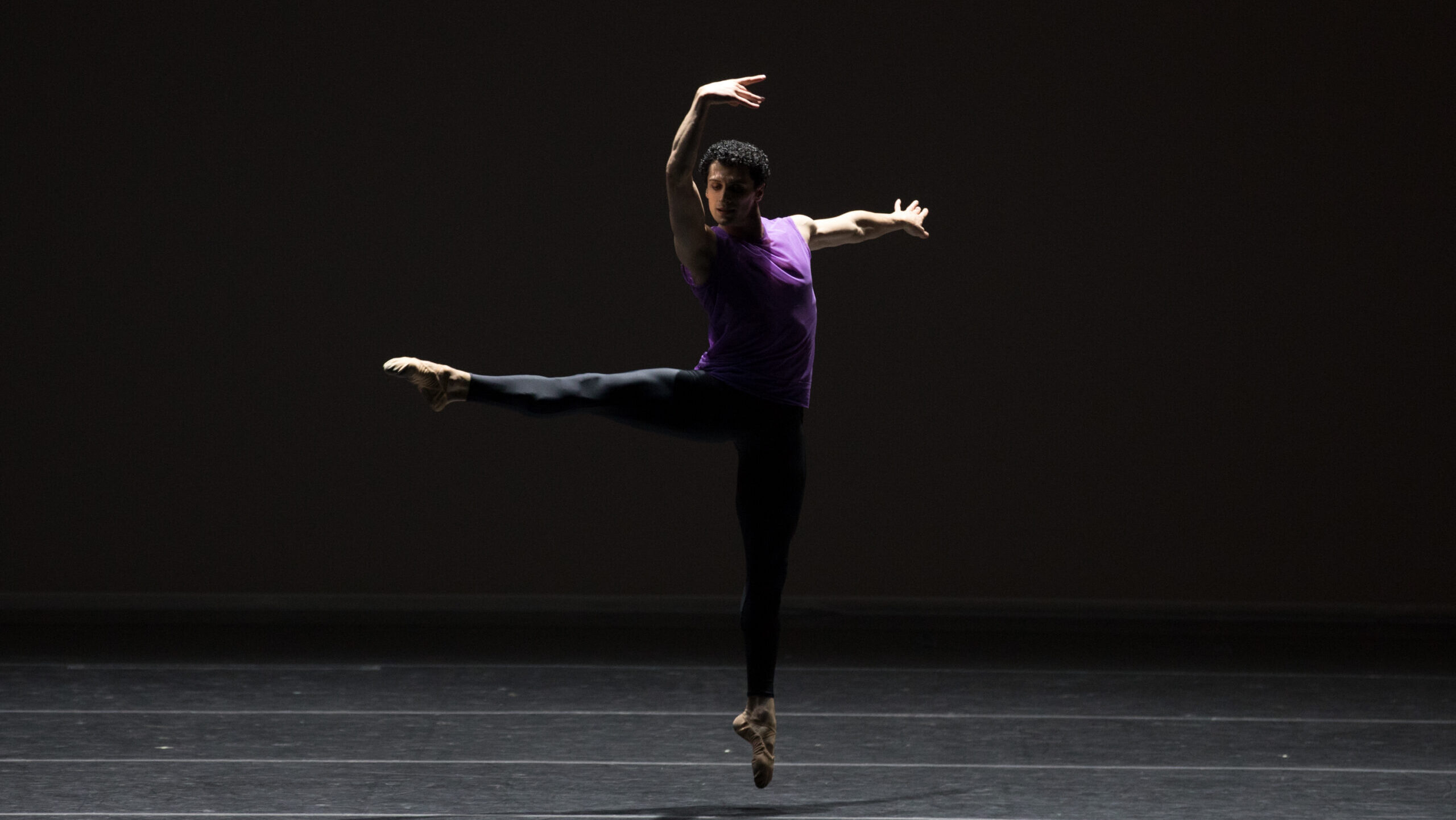 Lasha Khozashvili jumps with one leg extended 90 degrees a la seconde, his arms in high third with a slight twist in his shoulders. He wears black tights and a deep purple tank top.