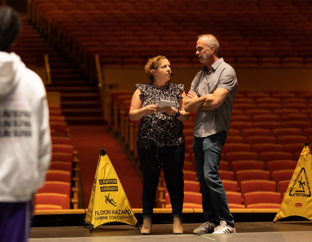 A woman holding a small notepad and pencil stands onstage and talks to a man on her left. They wear casual clothing and stand near yellow cones that say "Caution Floor Hazard." The house of the theater is in the background.