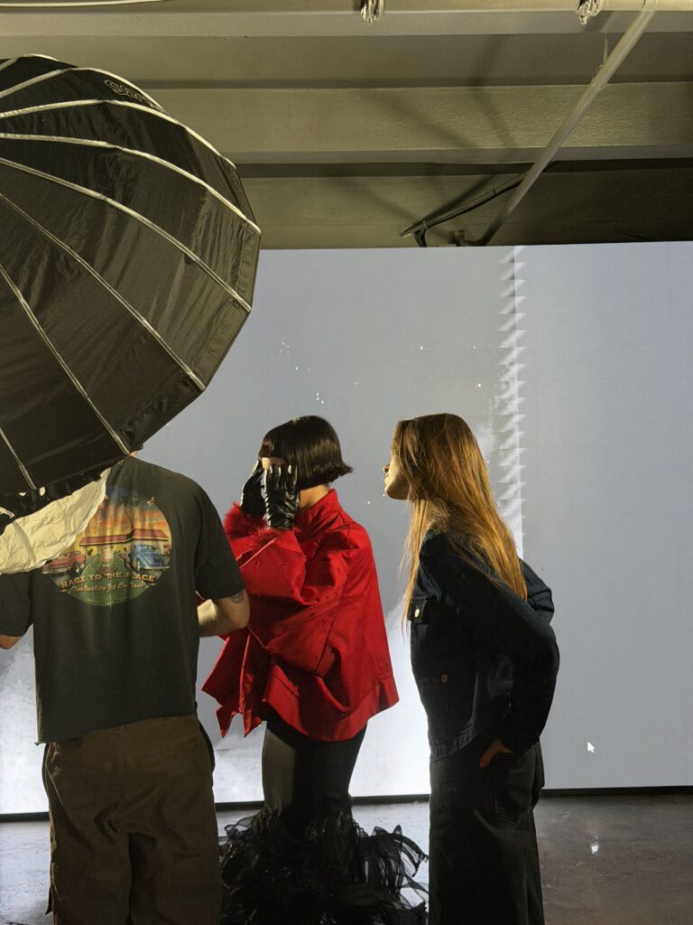 Cassidy Cock appears on the set of a photo shoot wearing black jeans and a black denim jacket. Next to her is a model wearing a bright red jacket and black gloves. A third person stands next to them wearing a dark grey t-shirt with a graphic on the back and shorts.