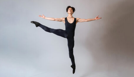 Takumi Miyake, wearing a black unitard and ballet slippers, jumps up with his right leg lifted in battement second. His arms are out to the side, palms up, and he lifts his head slightly.