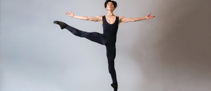 Takumi Miyake, wearing a black unitard and ballet slippers, jumps up with his right leg lifted in battement second. His arms are out to the side, palms up, and he lifts his head slightly.