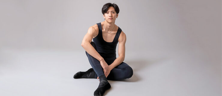 Takumi Miyake, wearing a black unitard and black ballet shoes, sits on the ground casually, hid left leg tucked under his right and his arms resting on his legs. He looks at the camera with a small smile.