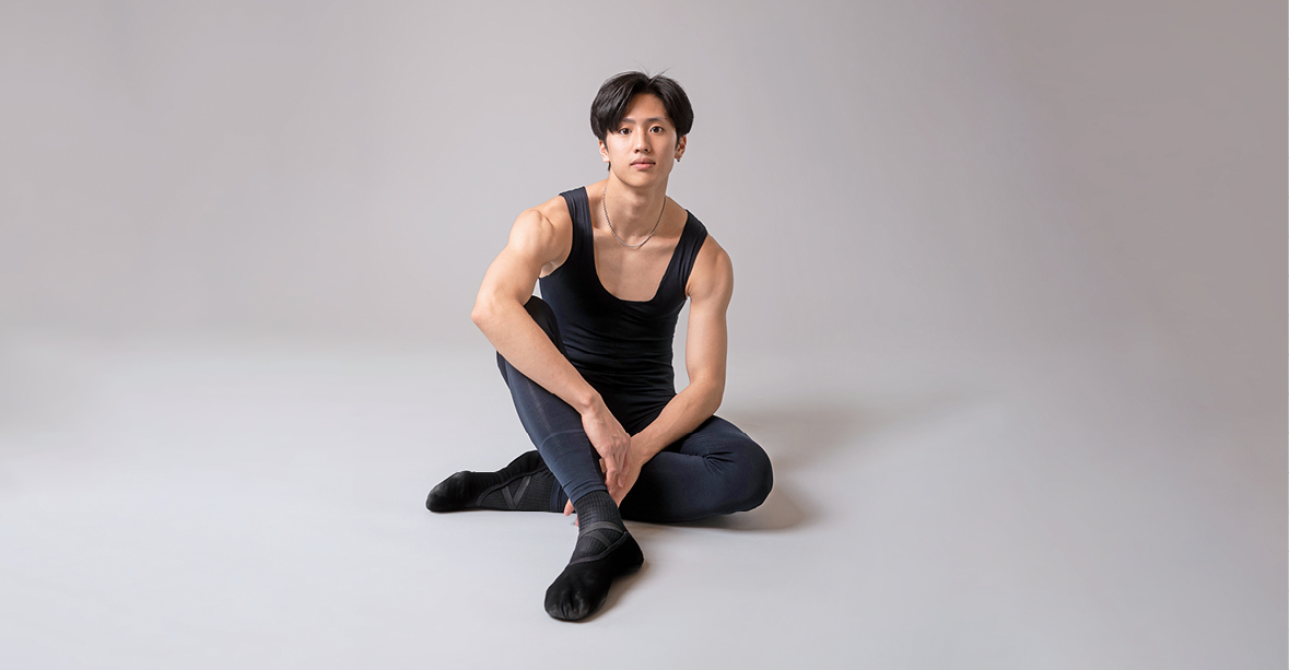 Takumi Miyake, wearing a black unitard and black ballet shoes, sits on the ground casually, hid left leg tucked under his right and his arms resting on his legs. He looks at the camera with a small smile.