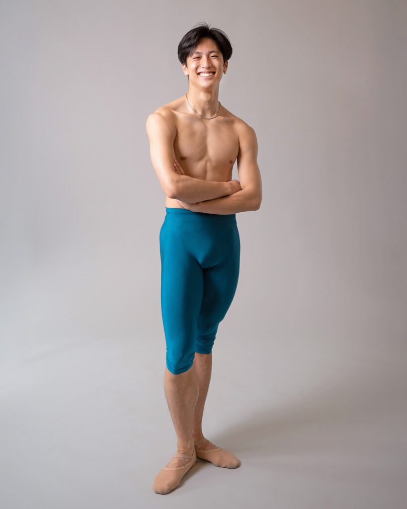 Takumi Miyake, wearing blue, knee-length tights and tan ballet slippers, stands casually with his arms crossed. He looks directly at the camera and smiles.