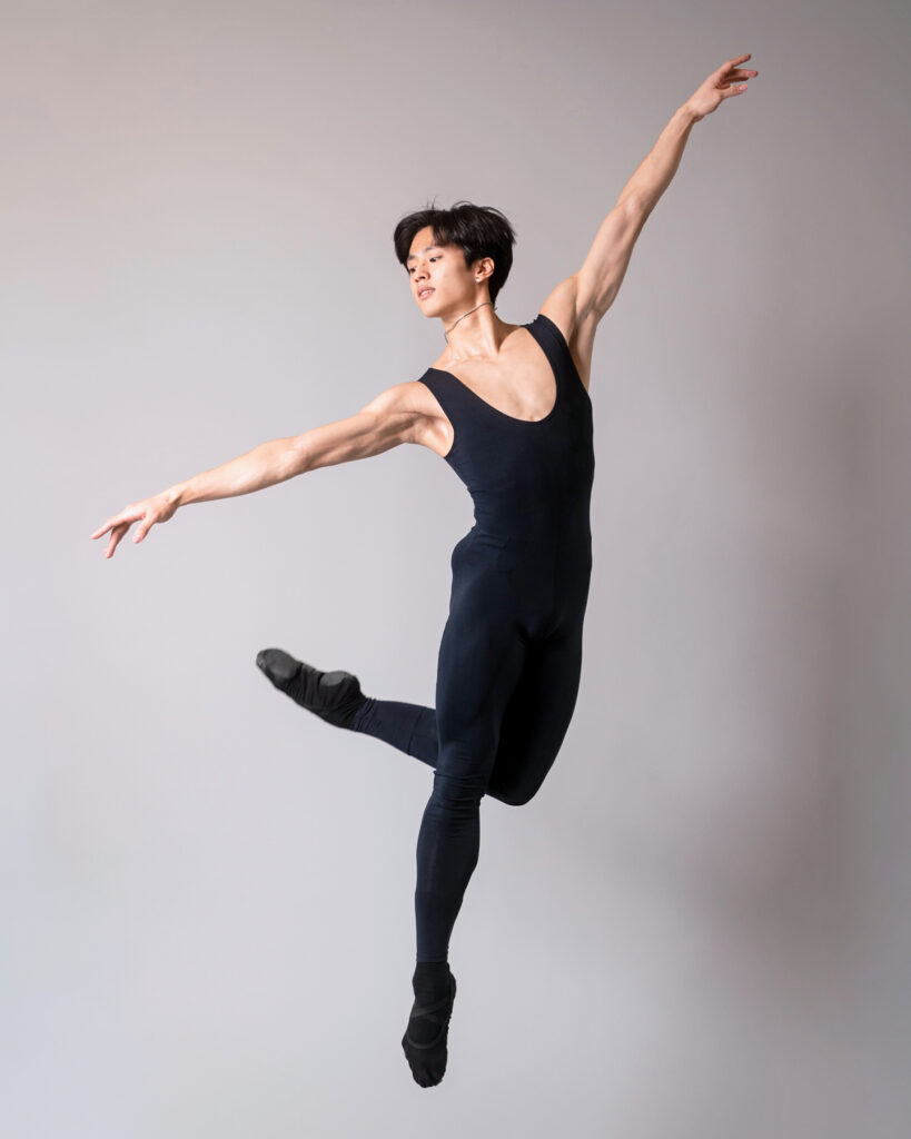Takumi Miyake, wearing a black unitard with black ballet slippers, jumps up, bending his left knee and crossing his leg behind his right with his arms held in a long diagonal. He looks over his right shoulder.