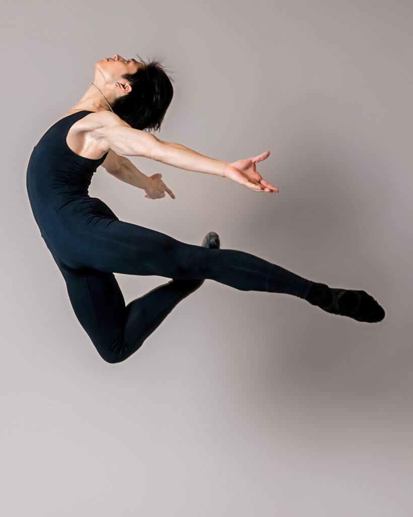 Takumi Miyake, wearing a black unitard and ballet slippers, jumps in profile facing his right. He throws his head back and presses his arms behind him, palms up. His left leg is in an arabesque line as he tucks his right leg underneath him.