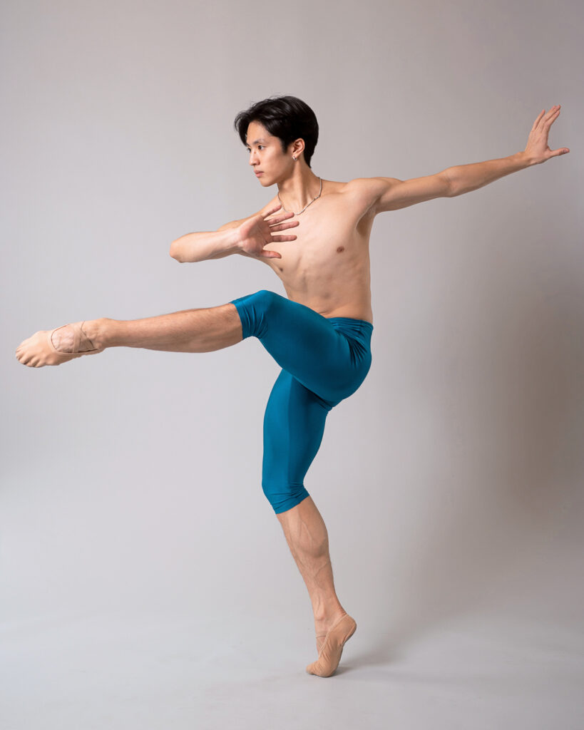 Takumi Miyake, wearing blue, knee-length tights and tan ballet slippers, does a piqué attitude devant in croisé with his left leg lifted. He pliés slightly on his right leg and presses his left arm back as he folds his right arm in towards his chest, palm facing out. His head is in profile, and he poses in front of a gray backdrop.