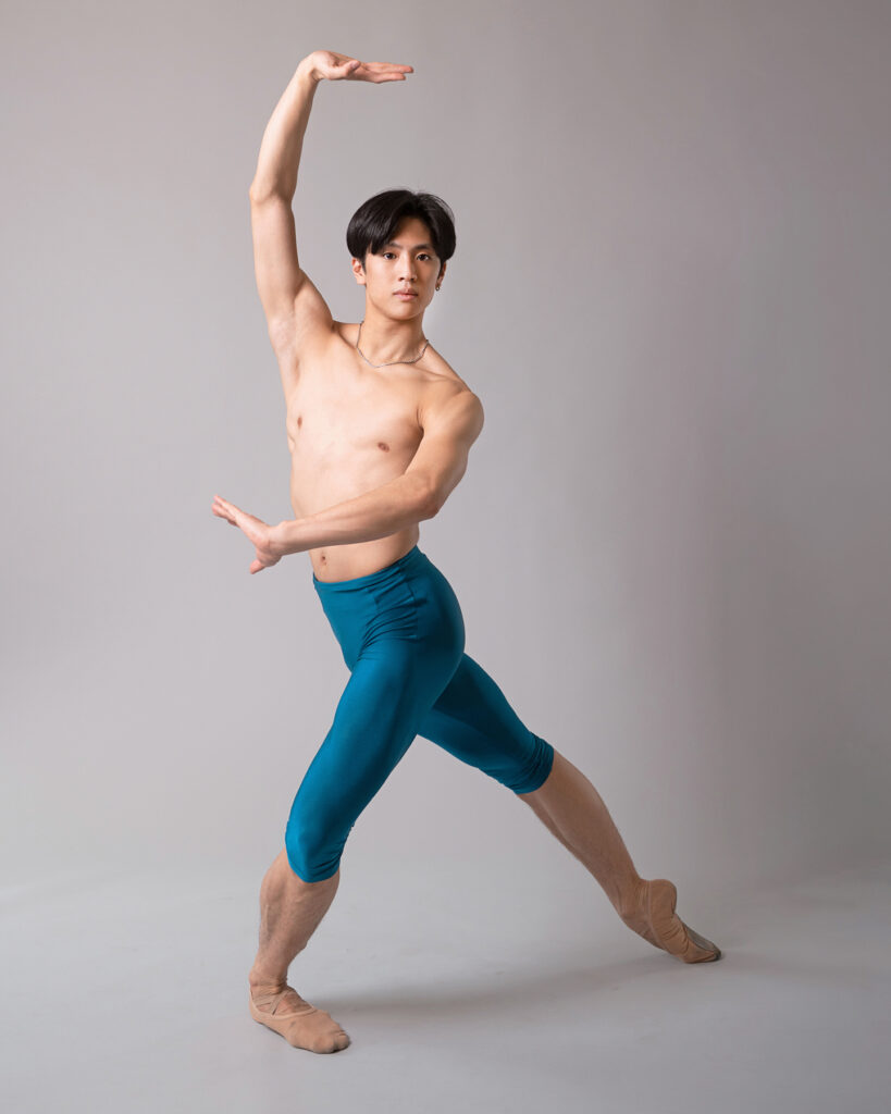 Takumi Miyake wears knee-length blue tights and poses in tendu croisé derriere. He cross his left arm in front of his torso and raises his right arm above his head, with his palms facing out, as he looks directly at the camera.
