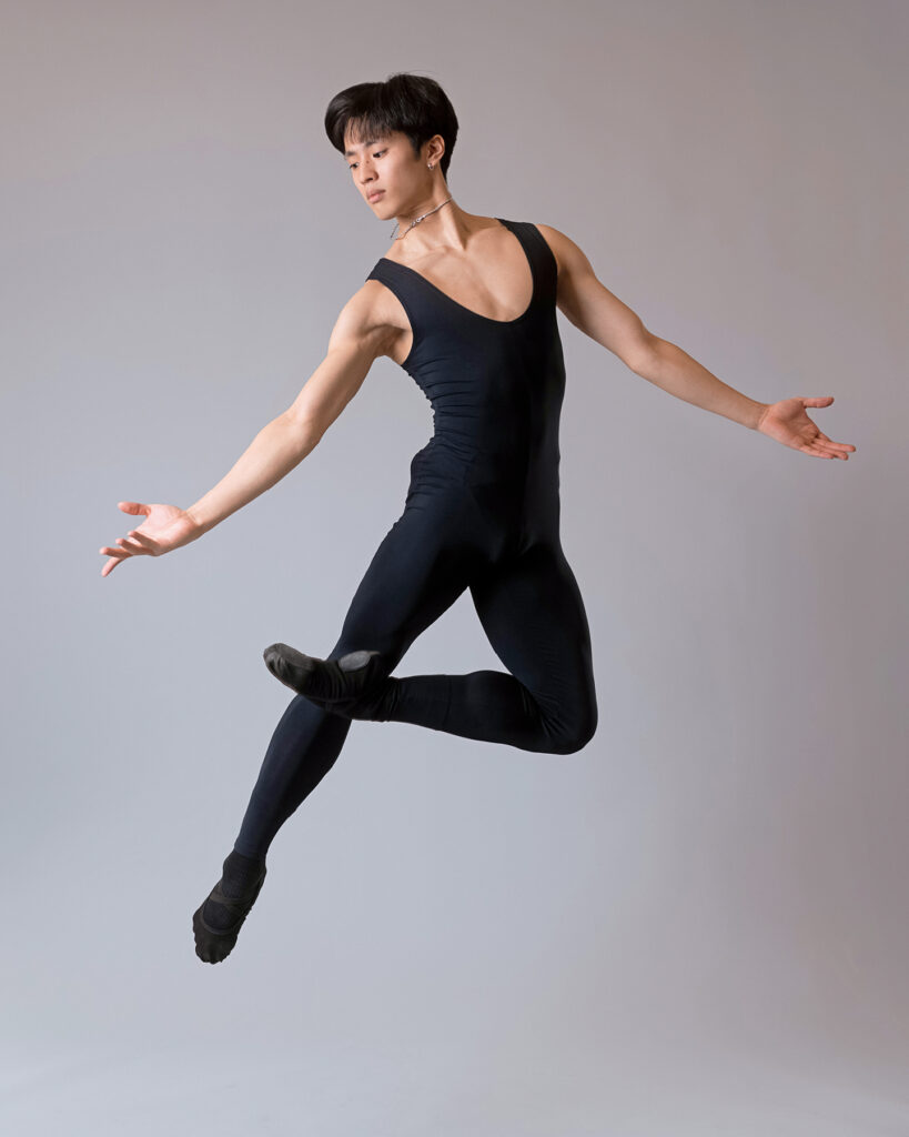 Takumi Miyake, wearing a black unitard and ballet slippers, jumps up with his left leg in passé, his foot slightly crosssing his right knee. He holds his arms low and out to the side, palms up, and looks over his right shoulder.
