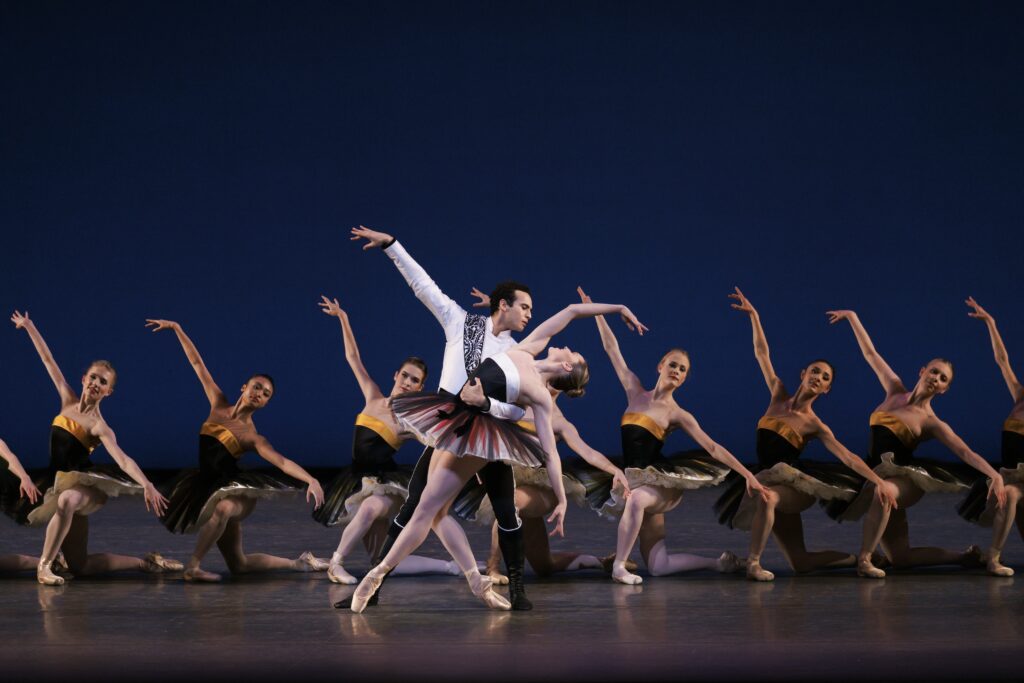 A man holds a woman by the waist as she arches back, her legs in fourth position on pointe with the left knee bent. She wears a white and black tutu with red trim; he is costumed in a white long-sleevedtunic with a black sash and black tights and boots. And corps of women in black and yellow tutus pose on their knee behind them.