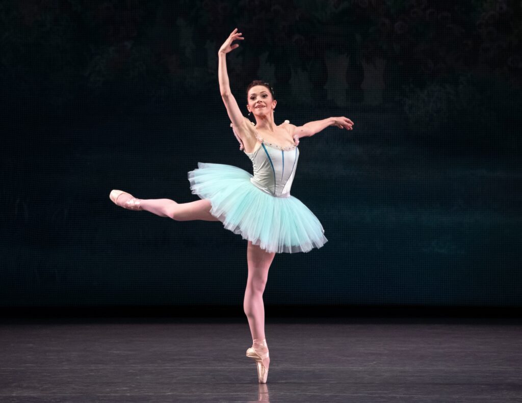 Megan Fairchild piqués in attitude derriere on pointe towards stage left with her right arm up and her left arm out to the side. She wears a light blue-green tutu and looks out towards the audience, smiling.