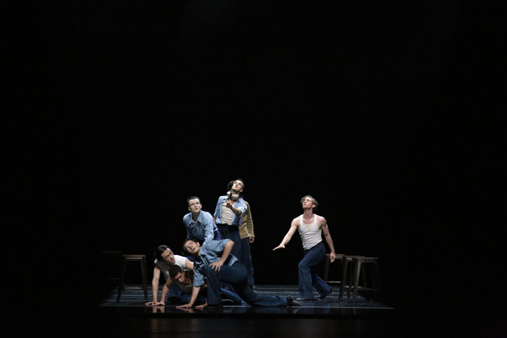 On a dark stage, a group of dancers dressed in 1940's–style clothing look up at the sky wearily, running toward the same spot where some dancers do a deep lunge.