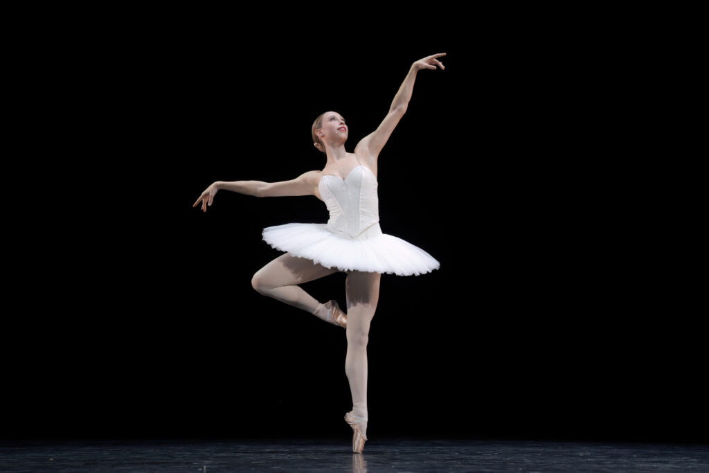 Calley Skalnik wears a white classical tutu and piqués in retiré with her foot crossed behind. She lifts her chest and looks up as her arms float lightly.
