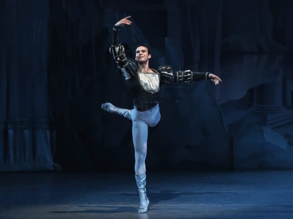 Ryan Tomash does an attitude derrierre with his left leg behind him and his right arm up. He wears a dark, medieval-style tunic with puffed sleeves, white tights, and white ballet boots.