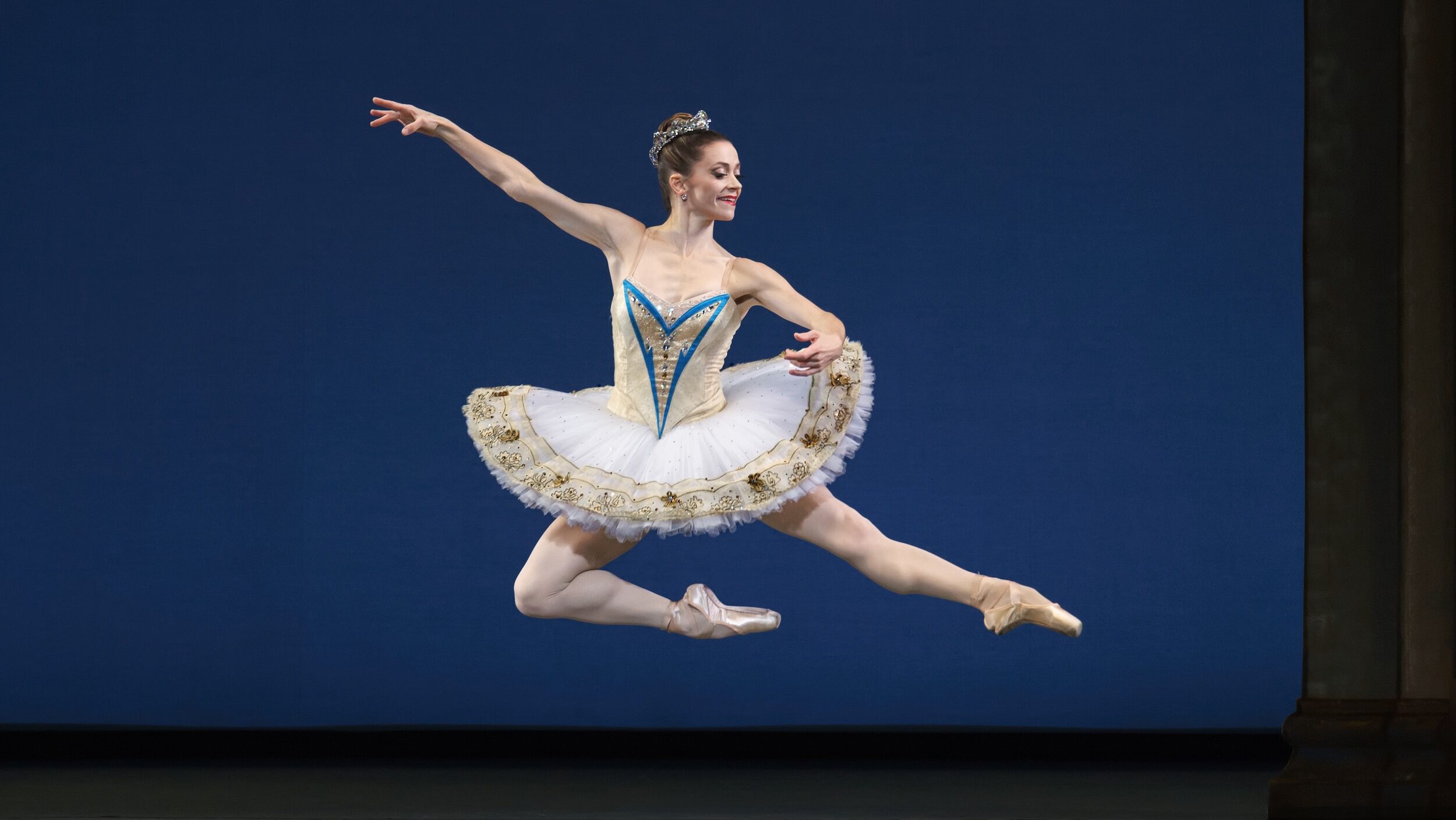 Megan Fairshild, wearing a white tutu with gold and bright blue trim and a sparkly tiara, does an Italian pas de chat towards stage left. She looks over her left arm with a smile.