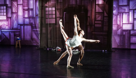 Onstage, two dancers perform a pas de deux in white shirts and shorts. Styles Dykes lunges forward and reaches up and forward to support his partner, Alexandra Troianos, who does an attitude penché in front of him. Behind them is a large purple and brown backdrop made to look like compartments carved in wood.