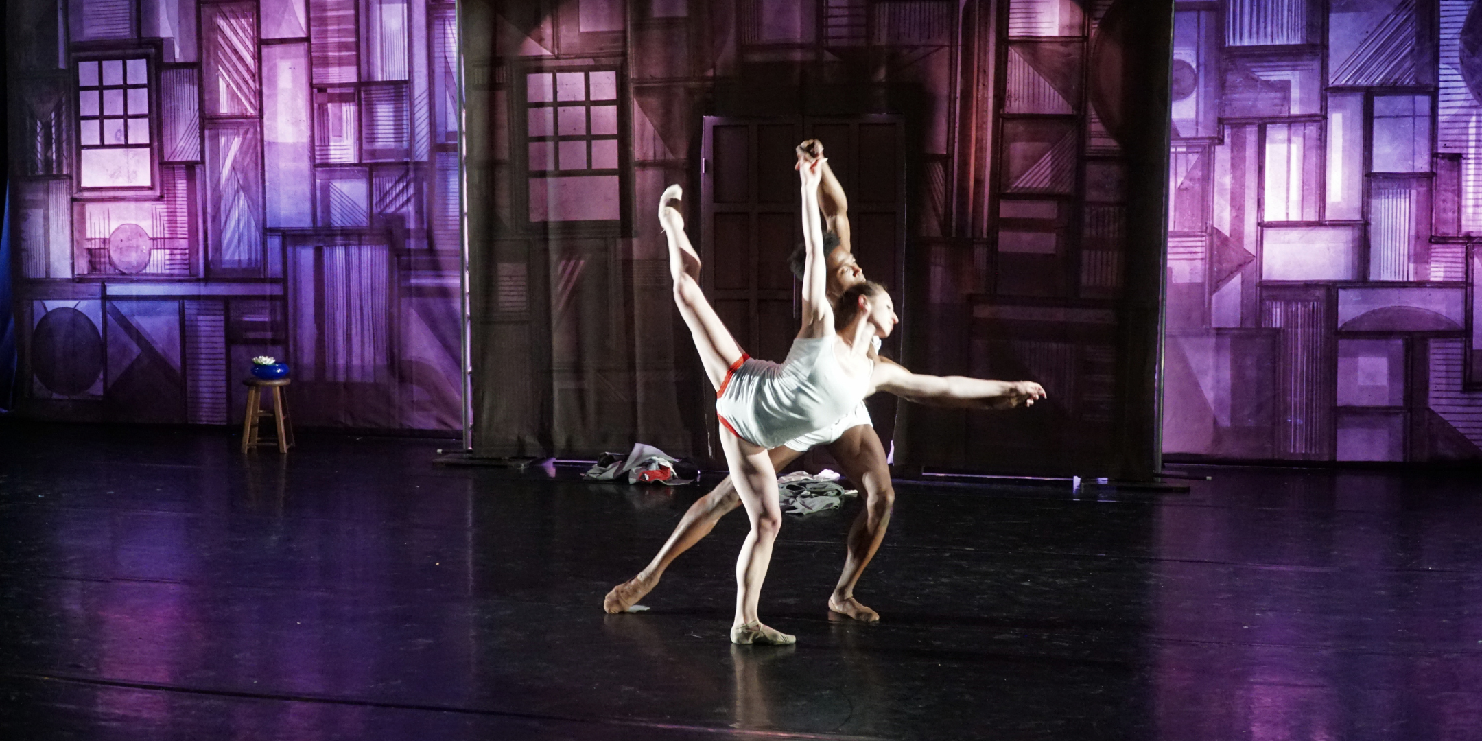 Onstage, two dancers perform a pas de deux in white shirts and shorts. Styles Dykes lunges forward and reaches up and forward to support his partner, Alexandra Troianos, who does an attitude penché in front of him. Behind them is a large purple and brown backdrop made to look like compartments carved in wood.