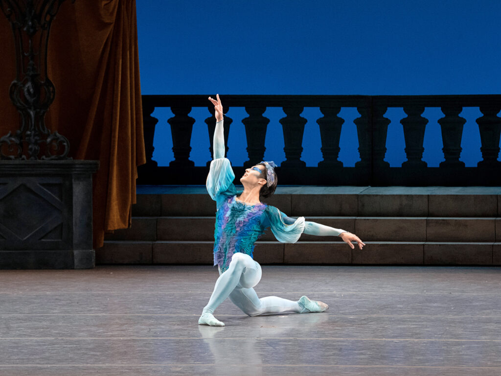Takumi Miyake kneels onto his right knee onstage and looks up at his lifted right arm, his other arm out to the side. He wears a billowy blue tunic with purple trim, blue tights and ballet shoes, and a blue headpiece.