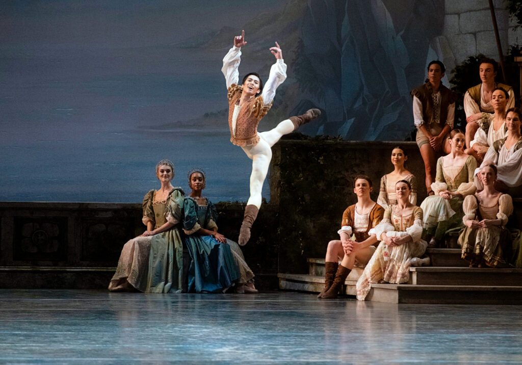 Takumi Miyake does a huge tour jeté, his arms in high fifth, a joyous expression on his face. He wears a white billowy shirt with a brown vest over it, white tights, and brow ballet boots. Behind him, a group of dancers in aristocratic costumes sit on a staircase and watch him.