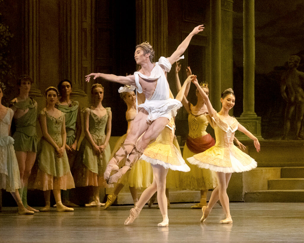 Takumi Miyake, wearing a short white toga-style tunic, tan ballet slippers with straps up the legs, and a wreath around his head, does an assemblé devant in effacé onstage. Two ballerinas in yellow tutus stand in B plus behind him, while a corps dancers light green and pink costumes stand and watch in the background.