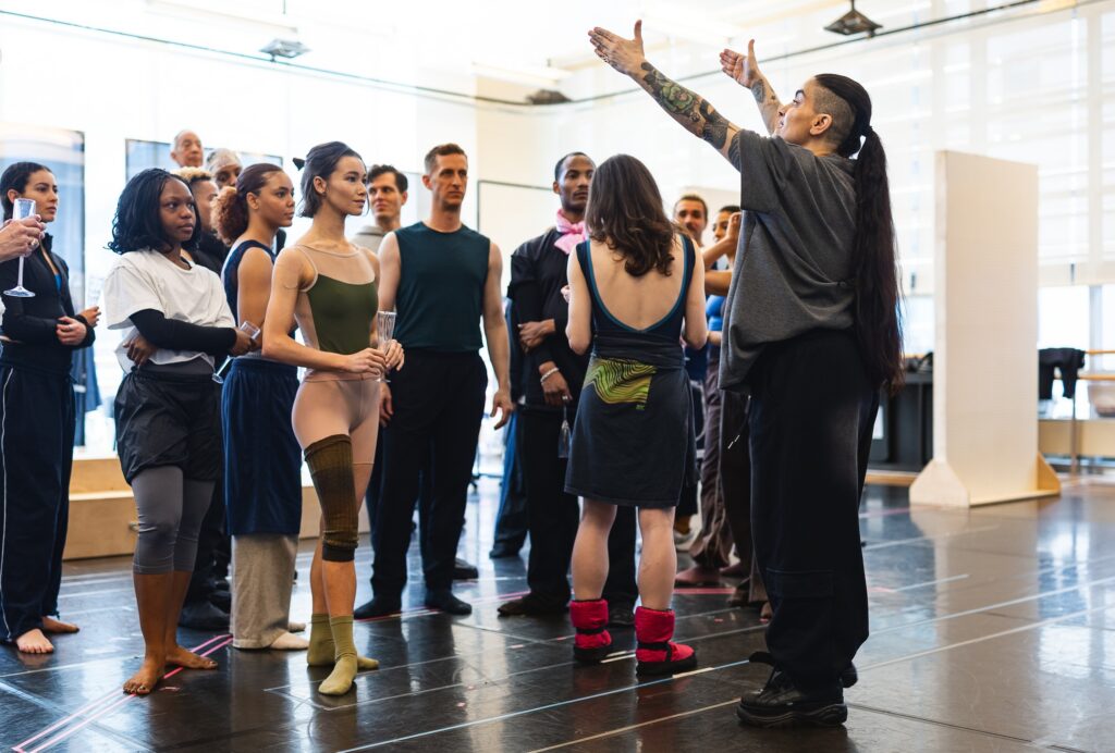 Sonya Tayeh stands with her arms up, wearing a grey shirt and black pants, giving notes. A group of dancers stands wearing athletic clothing, listening to her.