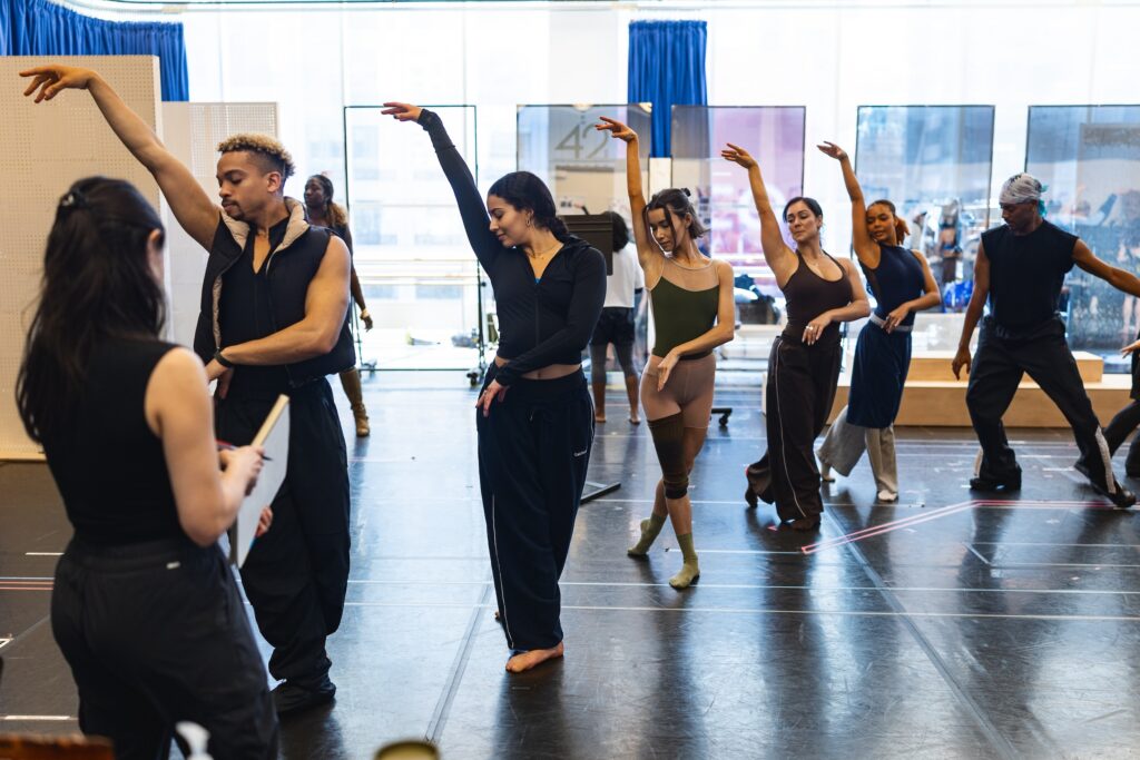 Five dancers stand on a diagonal in B plus with their left leg back. They each have their right arm up and left arm bent low across their torsos. At the front, an individual wearing all black watches them, holding a notebook and pen.