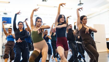 In a brightly lit studio, a large group of dancers wearing athletic clothing pose with their right arm up and left arm bent in front of them at chest height. They each step forward in plié with their right leg.