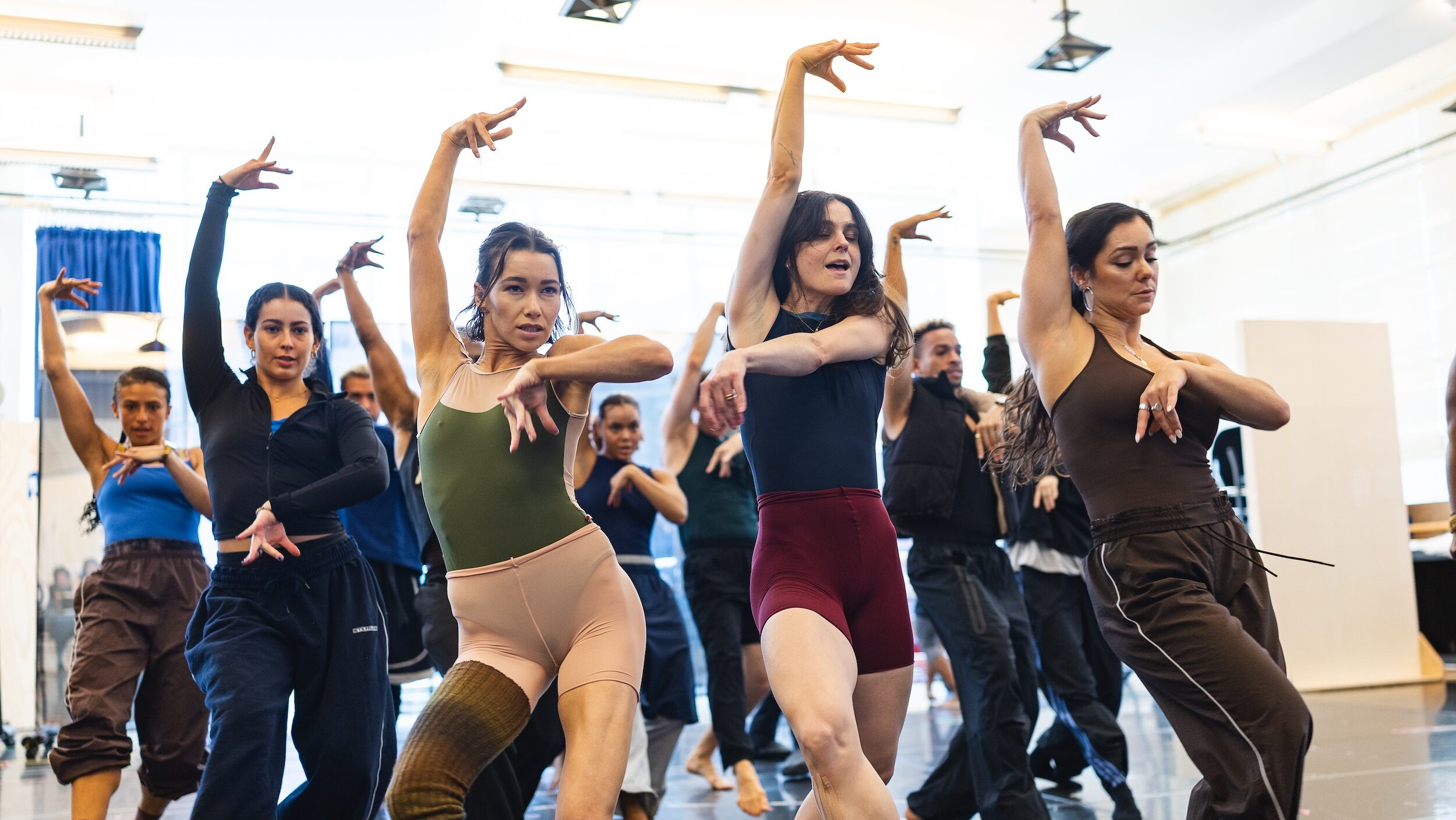 In a brightly lit studio, a large group of dancers wearing athletic clothing pose with their right arm up and left arm bent in front of them at chest height. They each step forward in plié with their right leg.