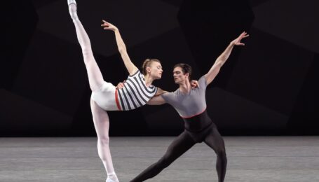 Miriam Miller and Peter Walker dance a pas de deux onstage. Miller, wearing a black and white striped leotard with a red waistband and and white tights, does a penché on pointe facing stage left as Walker lunges next to her, holding her waist. Miller put her arm around Walker's shoulder and lefts her right arm up. Walker wears a gray and black unitard.