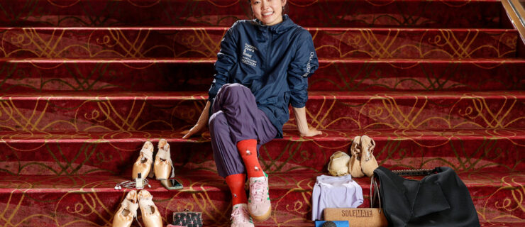 On red carpeted stairs, a ballet dancer sits with her legs crossed and leans on her hands behind her. She wears a warmup jacket and purple trashbag pants with red socks. Around her are the contents of her dance bag