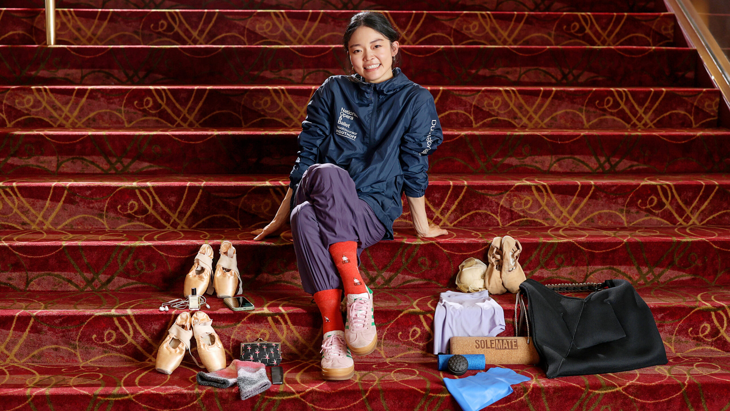 On red carpeted stairs, a ballet dancer sits with her legs crossed and leans on her hands behind her. She wears a warmup jacket and purple trashbag pants with red socks. Around her are the contents of her dance bag