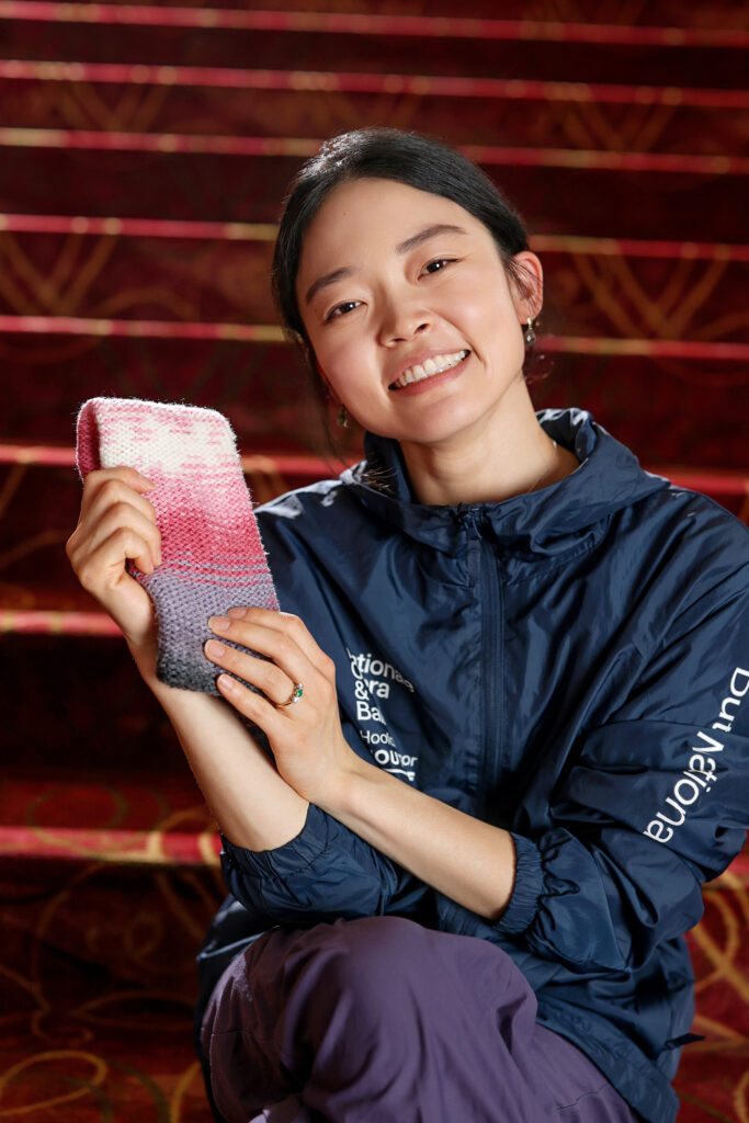 Riho Sakamoto holds one hand-knit legwarmer with a pink and gray pattern and smiles at the camera